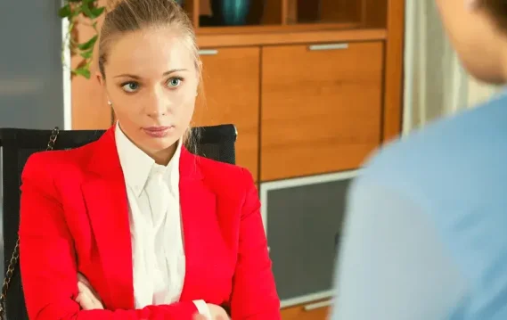 Woman in a red blazer with arms crossed, engaged in a serious conversation.
