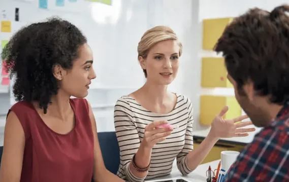 Three individuals engage in a discussion around a table, surrounded by colorful sticky notes on a wall, fostering a collaborative atmosphere.