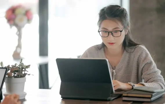 Woman wearing glasses focused on working at a laptop in a bright, modern workspace with books and a plant nearby.