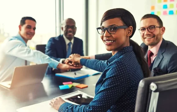 Smiling business professionals sitting around a table stack their hands in unity, showing teamwork and leadership.