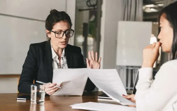 Two professionals in a business meeting discuss documents across a table with a glass of water and a smartphone, representing thoughtful decision-making for timing pay rate requests in job offers.