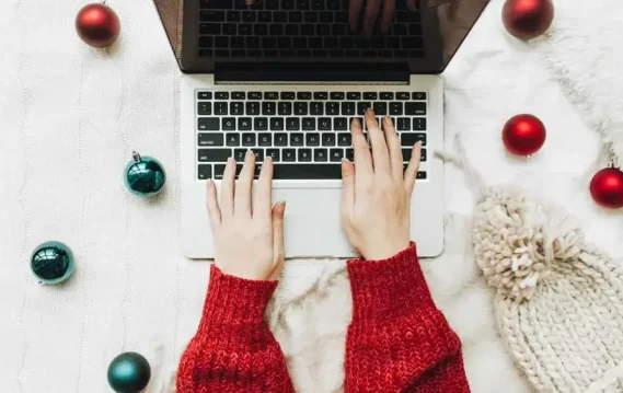 Person's hands typing on a laptop surrounded by festive ornaments and a cozy knit hat on a textured blanket.