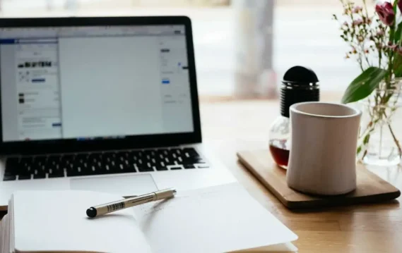 A cozy workspace setup with a laptop, an open notebook with a pen, a coffee mug, and a vase of fresh flowers on a wooden desk.