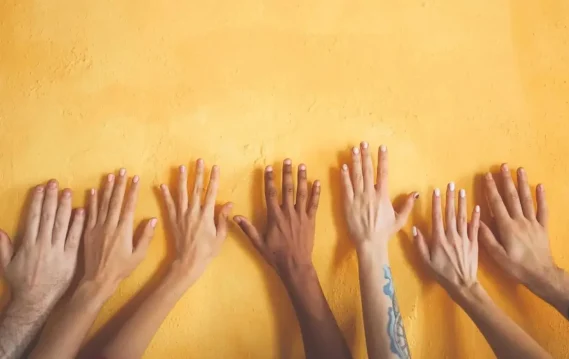 Diverse hands with different skin tones and nail designs against a yellow wall, symbolizing unity, diversity, and inclusion in the workspace.
