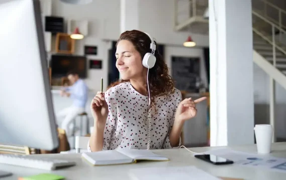 Music-Productivity-header Woman wearing headphones, smiling and dancing while working at her desk, enjoying music during work.