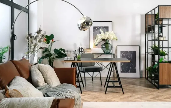 Stylish living space with a brown leather sofa, home office setup with a glass desk, and decorative plants in a light-filled room.