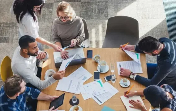 A group of people collaborates around a table, reviewing documents, laptops, and cups of coffee in a bright, modern workspace, promoting creativity.
