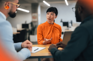 Three professionals discussing recruitment strategy in a modern office, with a woman speaking across a conference table.