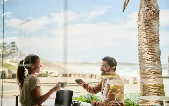 A woman and a man enjoying coffee at a seaside café with palm trees and ocean views, and a laptop on the table.