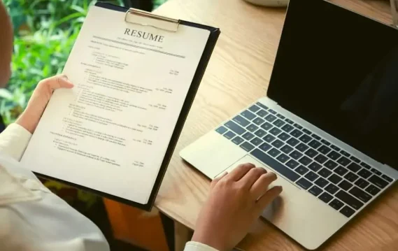 Person reviewing a printed resume on clipboard while using a laptop on a wooden desk.