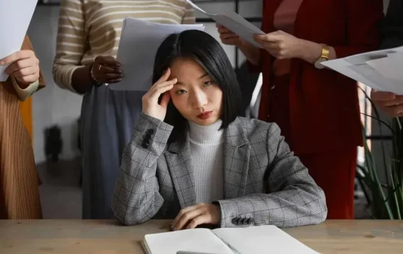 Stressed businesswoman surrounded by coworkers handing her documents, symbolizing workplace overwhelm