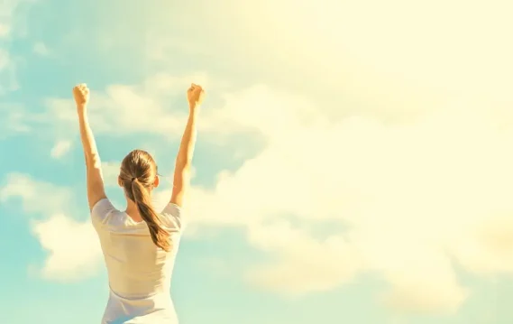 A woman stands with arms raised against a bright sky, symbolizing personal achievement, freedom, and a sense of triumph.