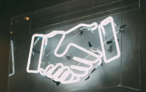 Virtual-networking-handshake A glowing neon sign of two hands shaking, symbolizing agreement and partnership, against a dark background.