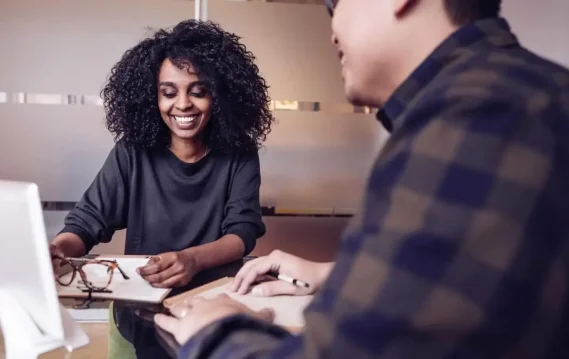 Woman with curly hair sitting at a desk, engaged in conversation, while another person writes notes in the foreground.