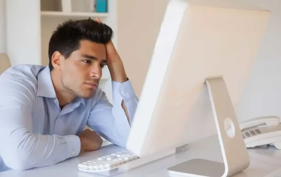 Frustrated office worker staring at a computer screen with a hand on his head.