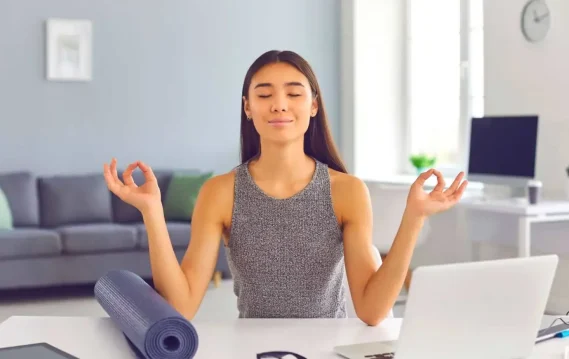 Woman meditating at her desk with eyes closed, a yoga mat beside her, and a laptop in front.