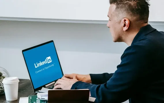Man working on a laptop displaying the LinkedIn logo, seated at a desk with papers and a coffee cup nearby.
