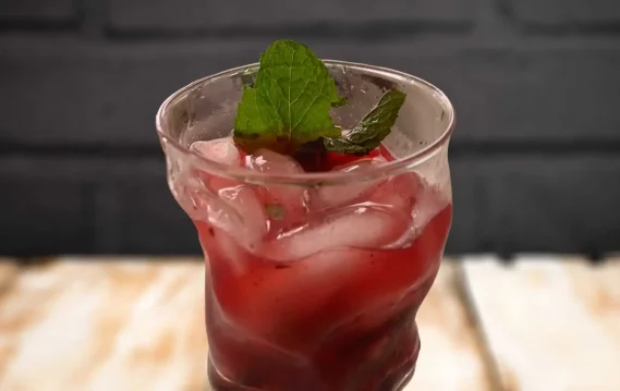 A refreshing red drink with ice and mint leaves, served in a curved glass against a dark background, representing a Bourbon Blackberry Smash.
