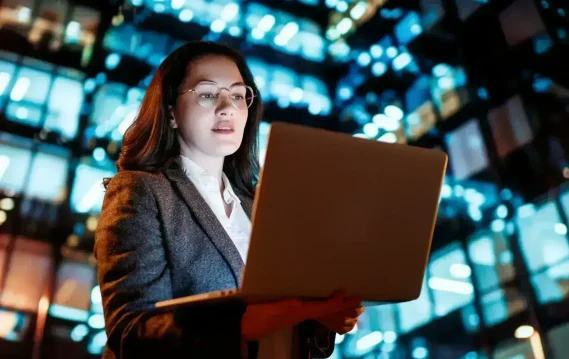 businesswoman-with-laptop-2024-11-05-07-43-09-utc-scaled Businesswoman holding laptop at night in front of illuminated city building, suggesting technology and modern work culture.