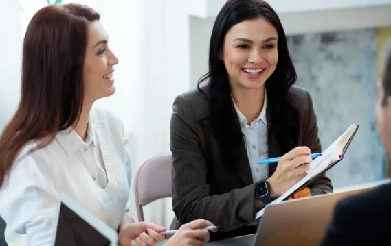Two women smiling and talking during a business meeting with notebooks and laptops.