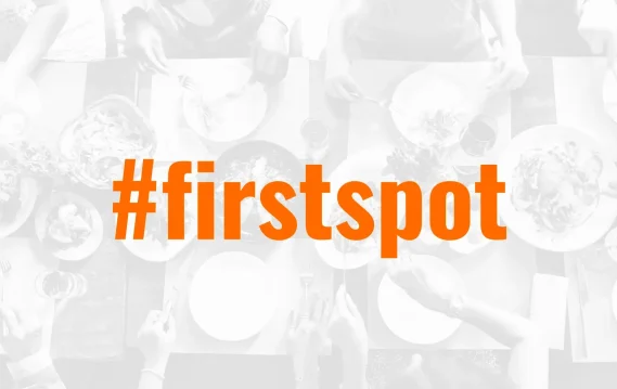 Aerial view of a dining table with hands reaching for food, overlaid with the hashtag #firstspot in bold orange letters.