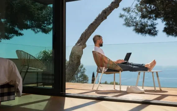 Person working on a laptop from a deck chair, overlooking the ocean and greenery, representing remote work in a scenic setting.