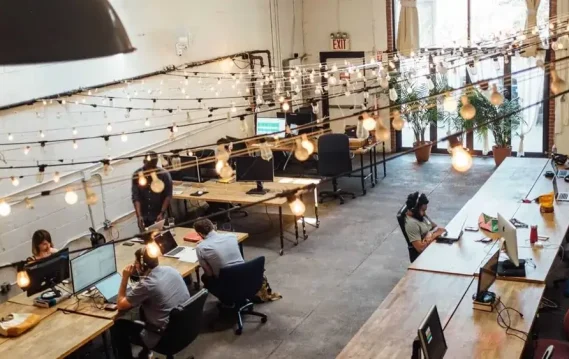 Open office space with people working at desks under hanging string lights.