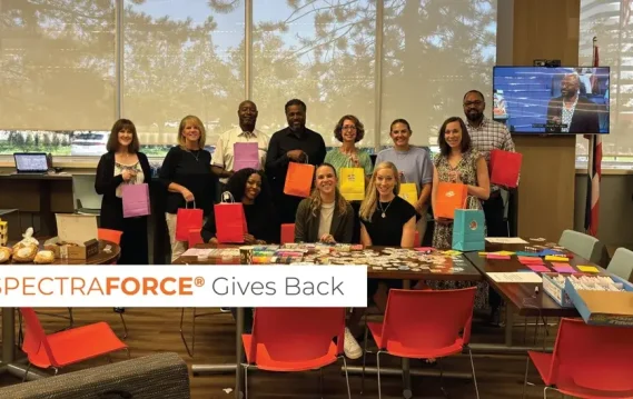image-7 A group of employees holds colorful gift bags in a bright office setting, with decorated tables and baked goods, symbolizing the charitable efforts of SPECTRAFORCE in support of Grace Centers of Hope.