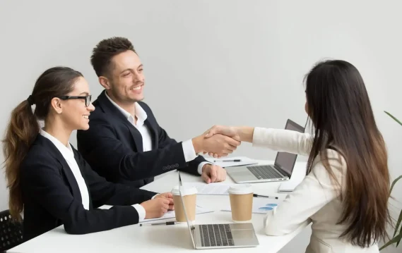 staffing-solutions-in-modern-day-scaled A woman in a white blazer shakes hands with a male interviewer while a female interviewer looks on, all smiling during a job interview.