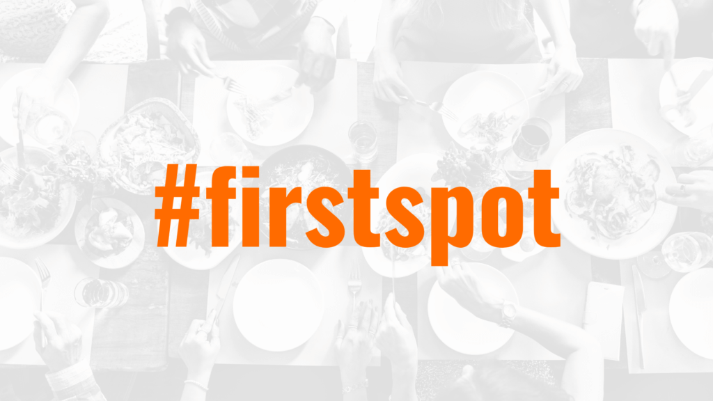 A table set with various dishes and utensils, featuring several people dining together. Overlaid in the center is the hashtag "#firstspot" in large, bold orange text. The image has a high-contrast black-and-white filter, except for the orange text.