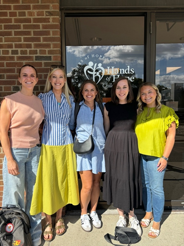 Five people stand smiling in front of a building entrance with a glass door displaying "Hands of Hope." They are dressed in casual summer clothes, and the brick wall and doorway are visible behind them. A backpack rests on the ground to the left.