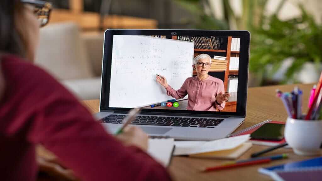 A person participates in an online lesson using their laptop. The laptop screen shows a teacher explaining mathematical equations on a whiteboard. The person is taking notes at a desk scattered with stationery and notebooks.