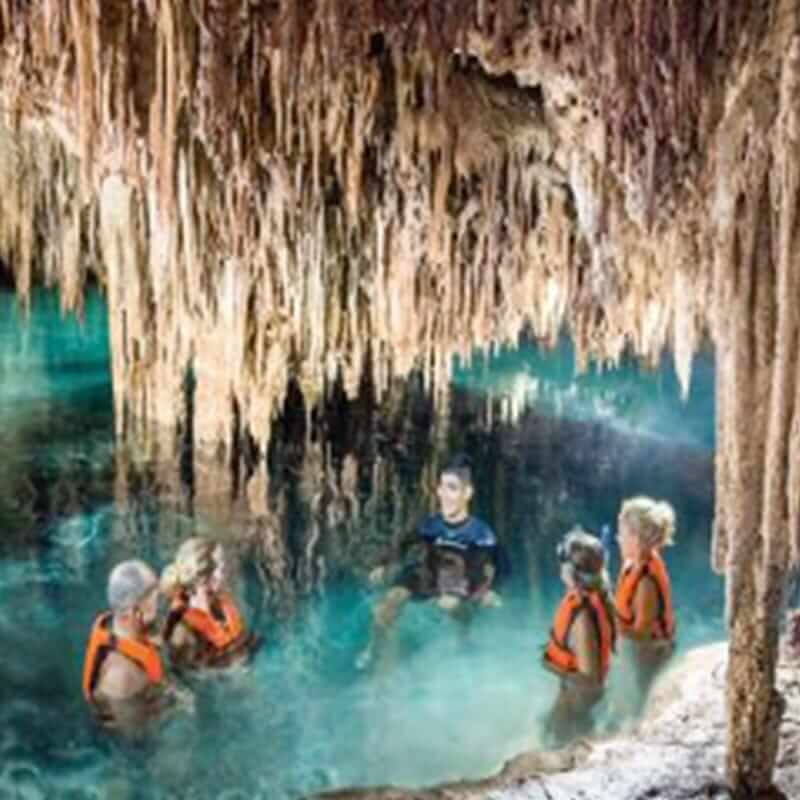 A group of five people wearing life vests explore a cave filled with water. They are surrounded by numerous stalactites hanging from the ceiling of the cave. The water is clear, and the lighting highlights the textured formations above and around them.